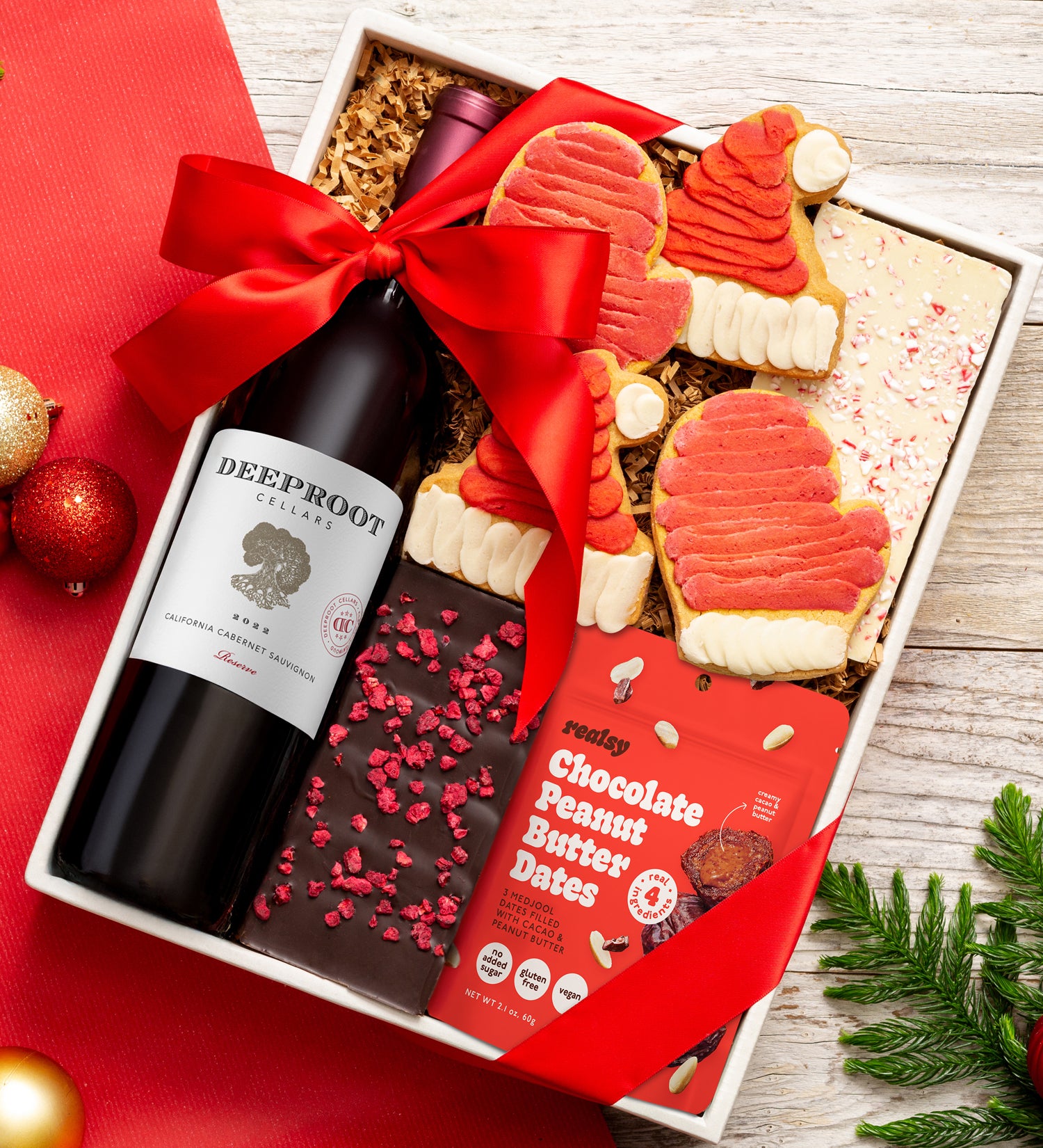 Gift box with wine, cookies, and chocolate peanut butter dates on a red surface.