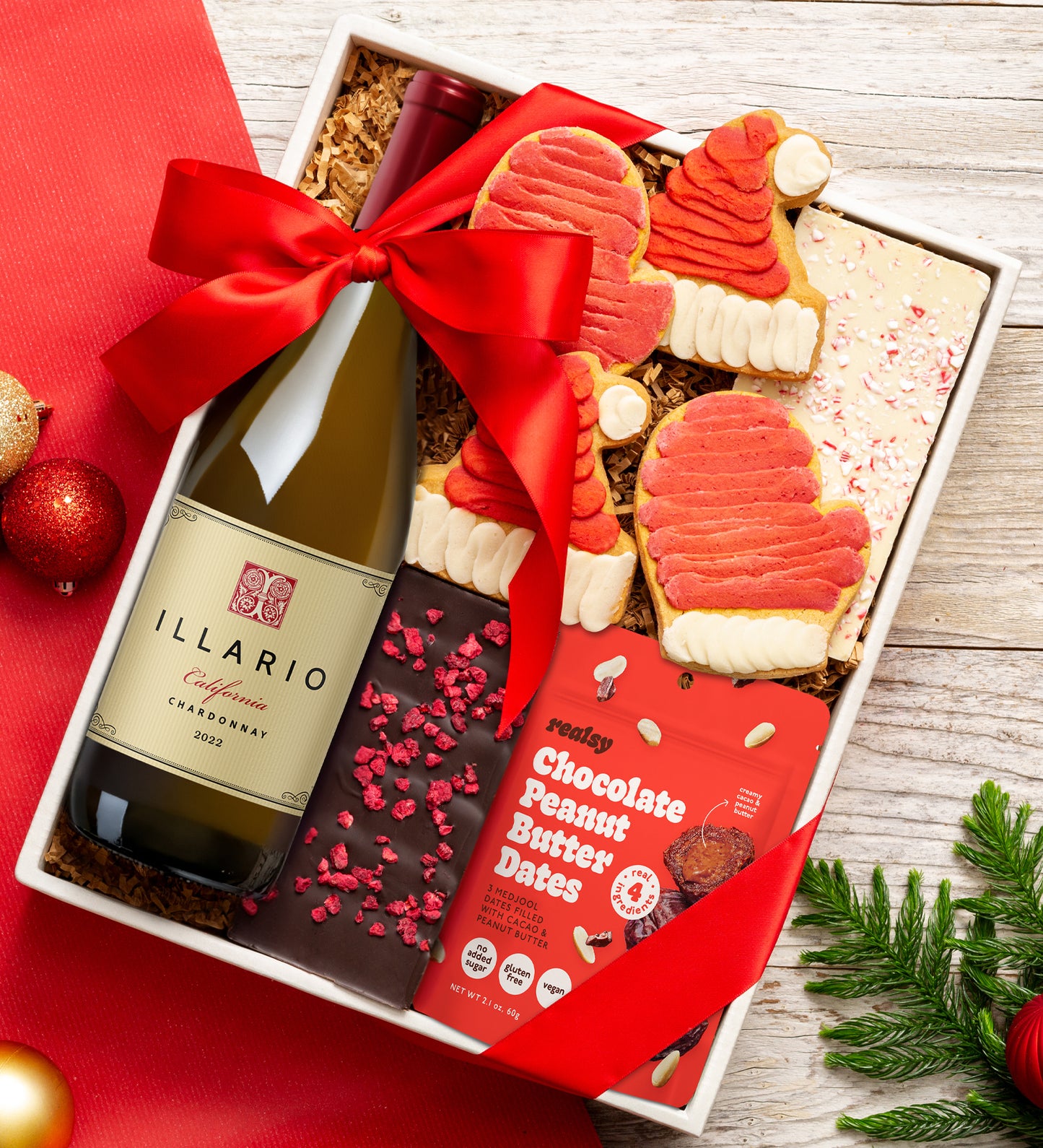 Gift box with wine, cookies, and chocolate peanut butter dates on a red surface.