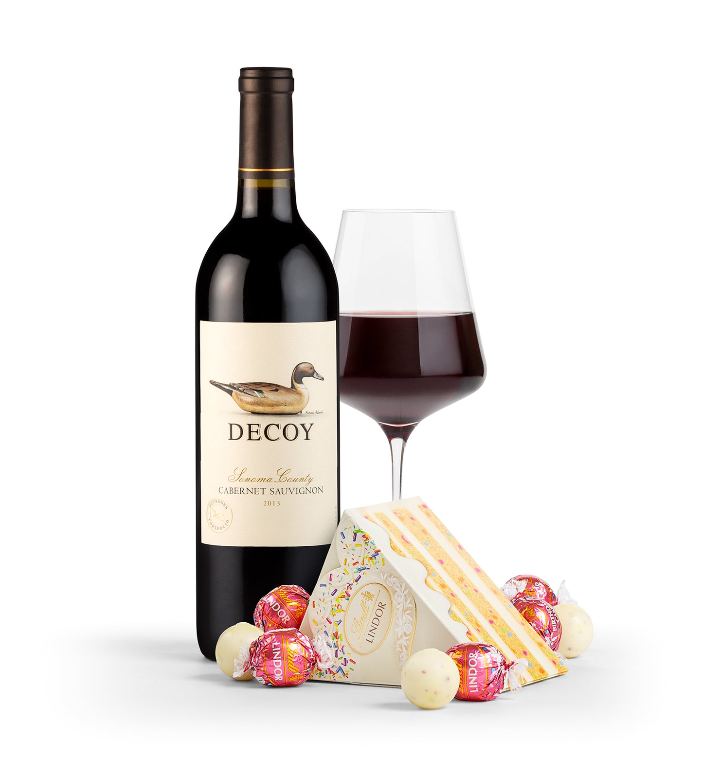 A bottle of Decoy Cabernet Sauvignon wine alongside a glass of red wine and a box of LINDOR Birthday Cake Truffles. The truffles box is open, displaying various funfetti-inspired white chocolate truffles.