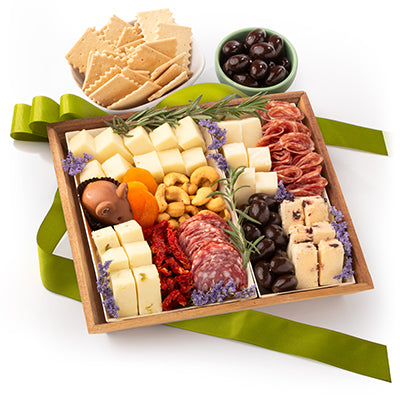 A gourmet charcuterie board gift box filled with cheeses, cured meats, nuts, dried fruit, crackers, and more, arranged in a wooden tray with a green ribbon bow on a white background.