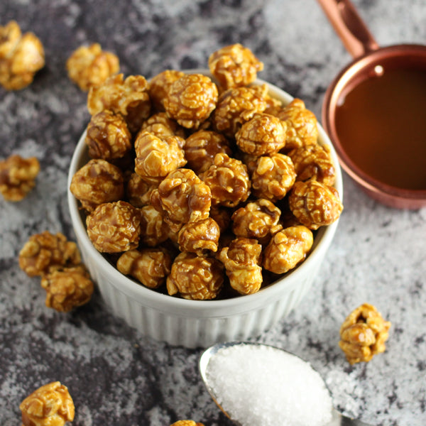Caramel popcorn in a white bowl on a gray surface with a small copper cup.