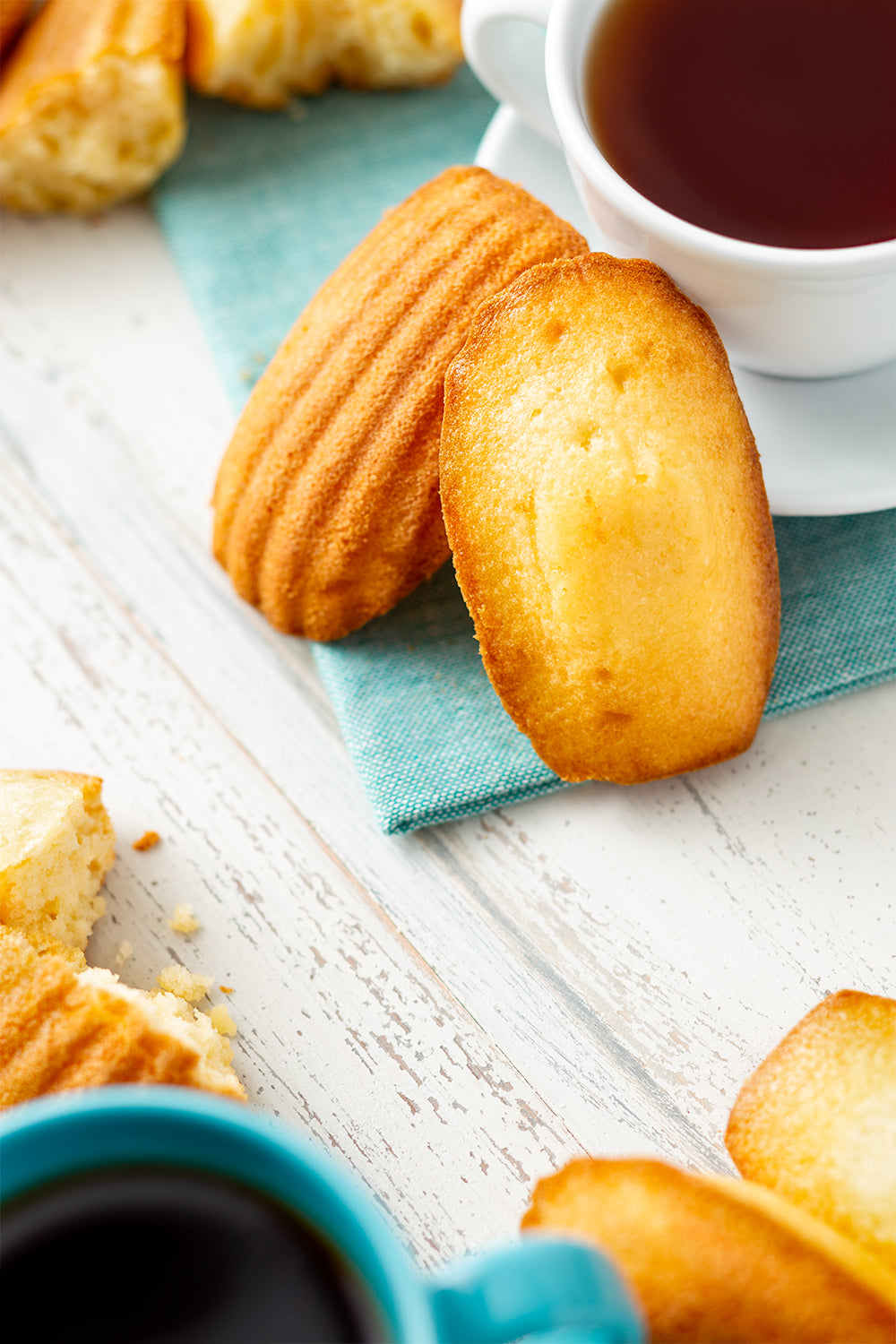 Madeleines on a turquoise napkin with a cup of coffee on a white wooden surface
