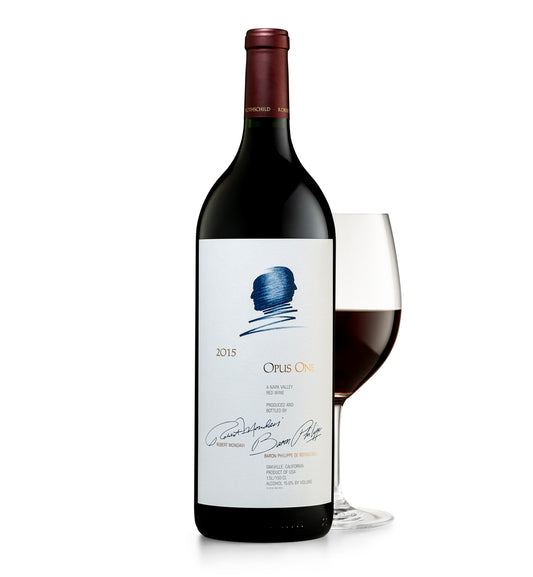 A magnum of Opus One wine, with a wine glass next to it. The bottle is upright and has a label with blue and red text, and a signature at the bottom.