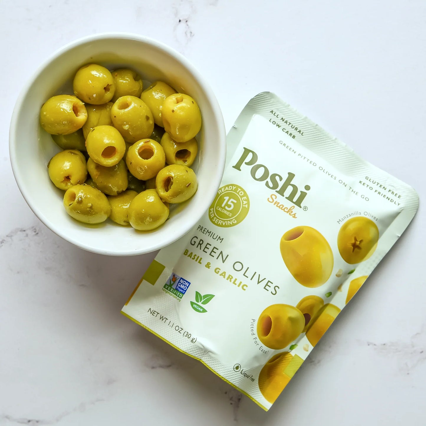 White bowl filled with green olives next to a Poshi Snacks package on a white background