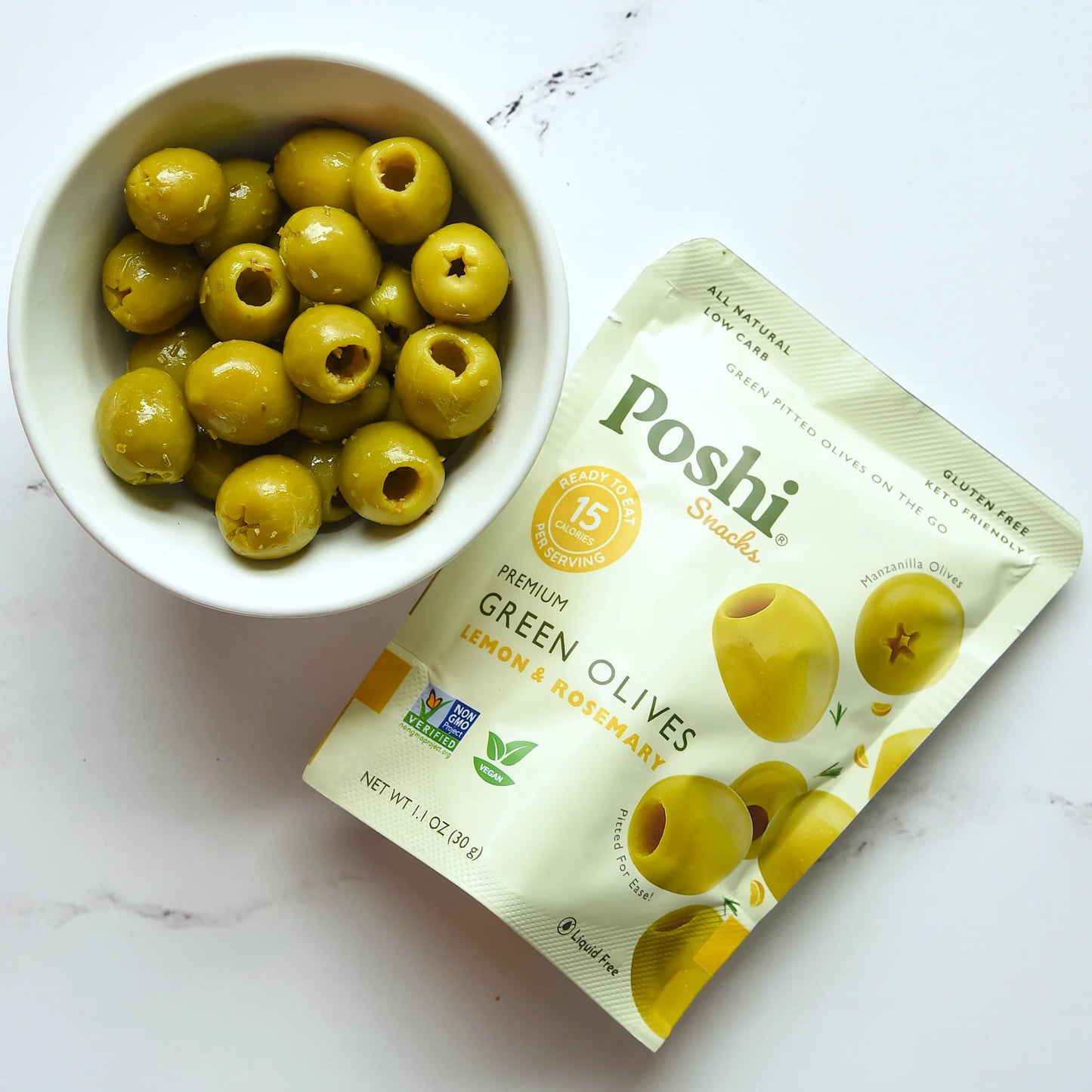 White bowl filled with green olives next to a Poshii Snacks package on a white marble surface