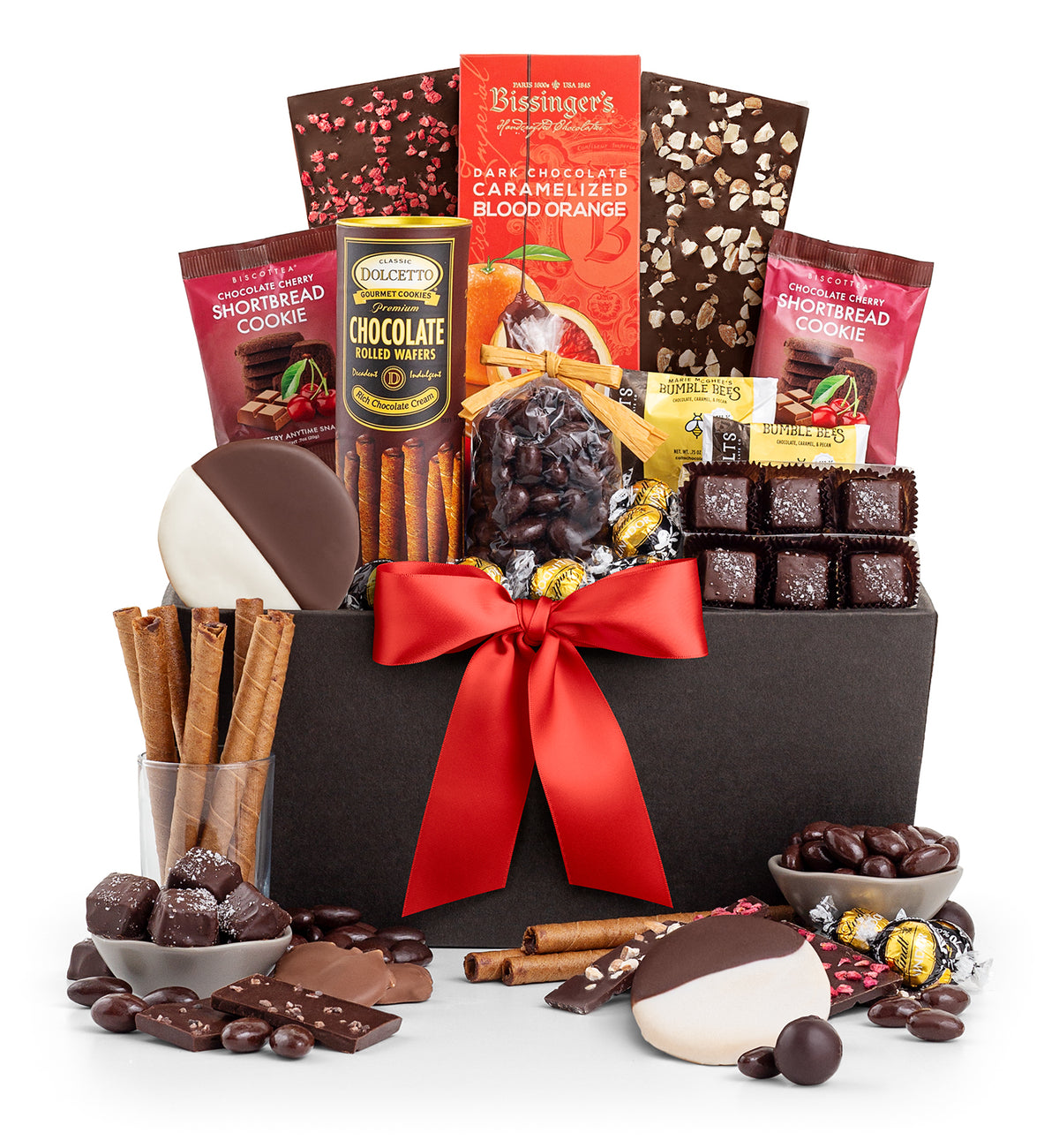 A chocolate gift basket filled with assorted chocolates, cookies, candy canes, cinnamon sticks, and wrapped treats, arranged in a black box with a large red ribbon on a white background.