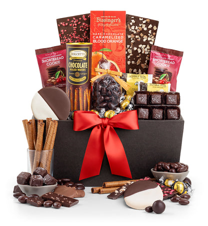 A chocolate gift basket filled with assorted chocolates, cookies, candy canes, cinnamon sticks, and wrapped treats, arranged in a black box with a large red ribbon on a white background.