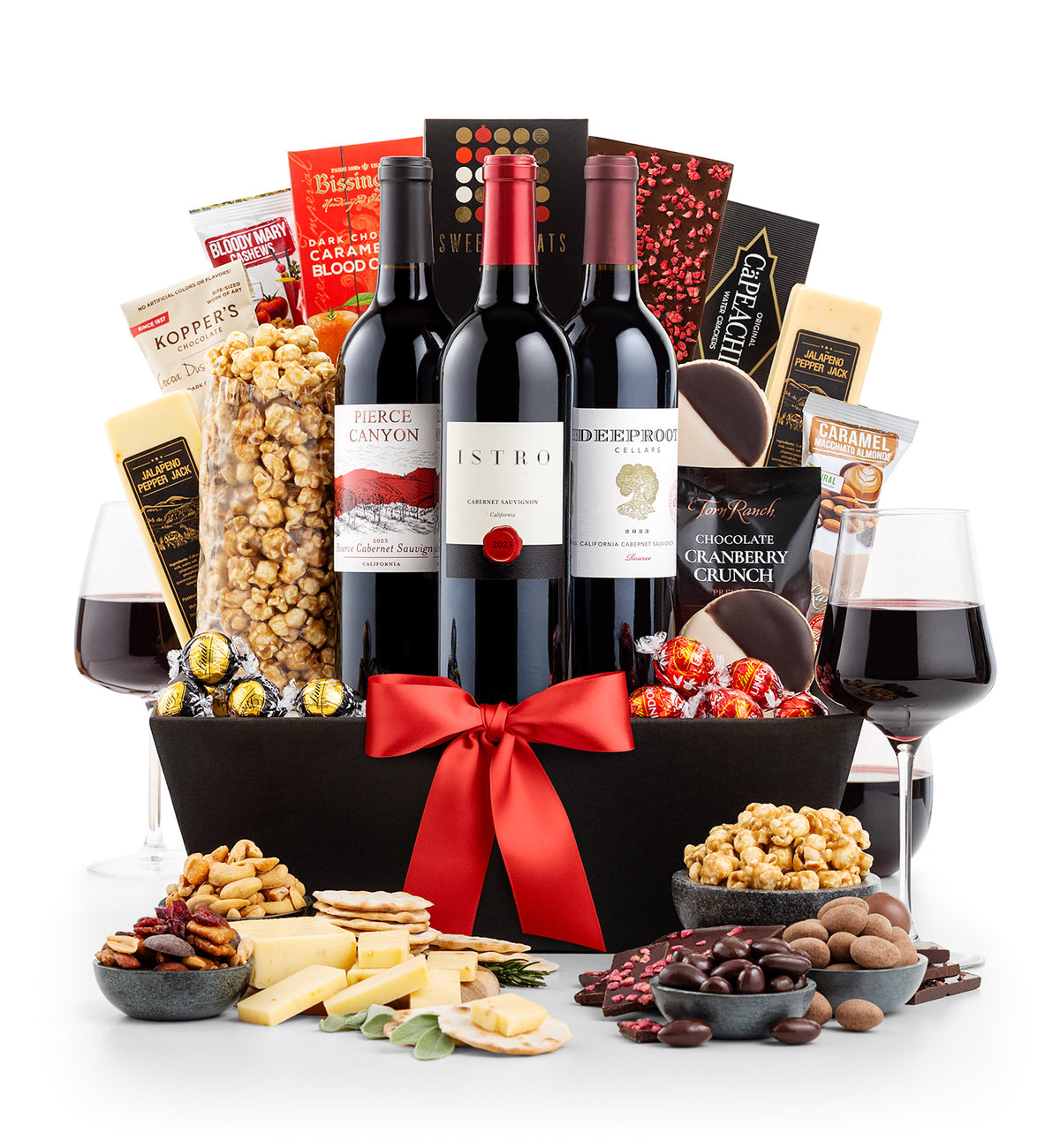 A gourmet wine and snack gift basket arranged in a black basket with a large red bow, featuring three red wine bottles, two glasses of red wine, assorted cheeses, nuts, crackers, chocolates, and packaged snacks on a white background.