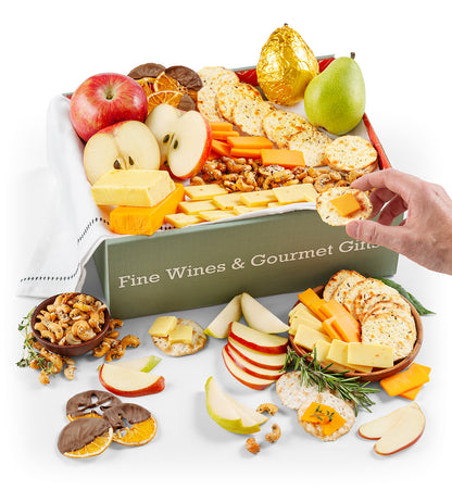 Gourmet gift box with hand reaching in, fruits, cheese, and crackers on a white background.