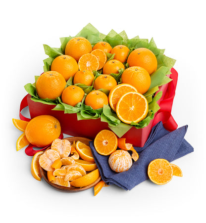 A gift box of fresh oranges and mandarins arranged in a red box lined with green tissue paper, with a red ribbon, a small wooden bowl of peeled mandarins, and a navy blue napkin on a white background.
