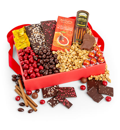 A red gift box filled with assorted chocolates, candy-coated chocolate beans, caramel popcorn, cinnamon sticks, and wrapped candies, arranged on a white background with some items spilled around the box.