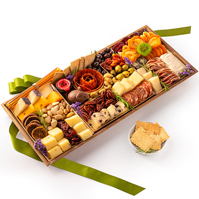 A gourmet charcuterie and cheese gift board arranged in a wooden tray, featuring various cheeses, cured meats, crackers, nuts, olives, dried fruit, and citrus slices, with a green ribbon and a small side dish of crackers on a white background.