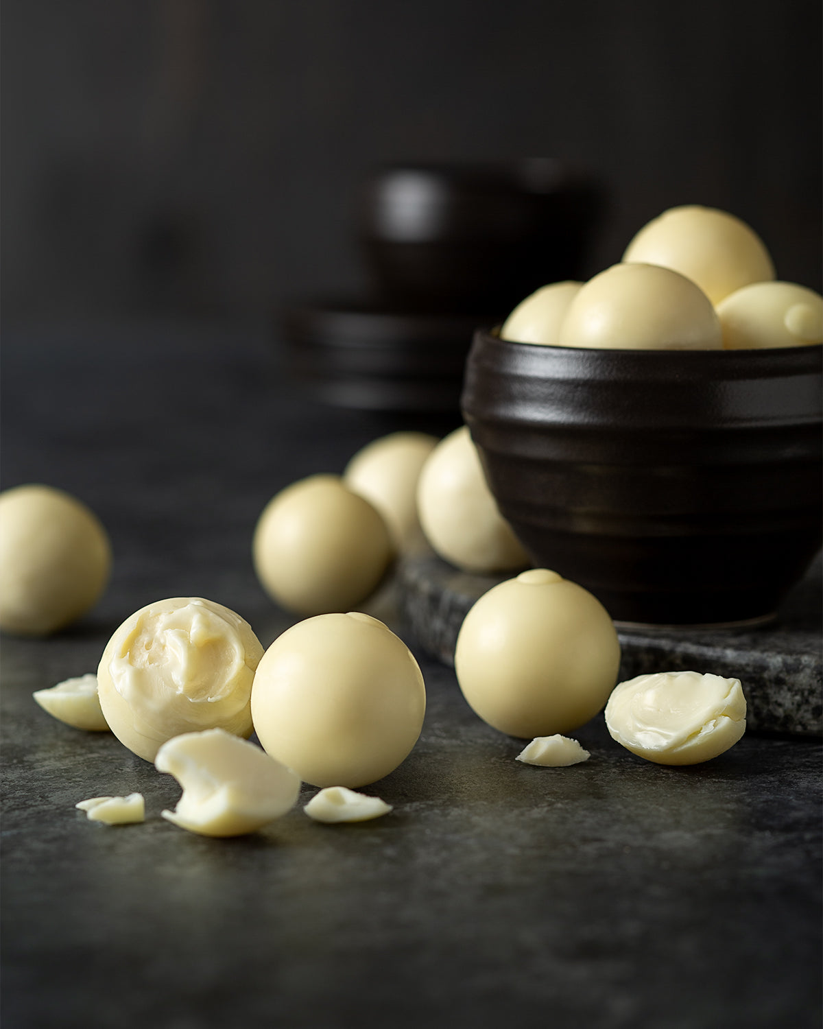 White chocolate truffles on a dark surface with a bowl in the background