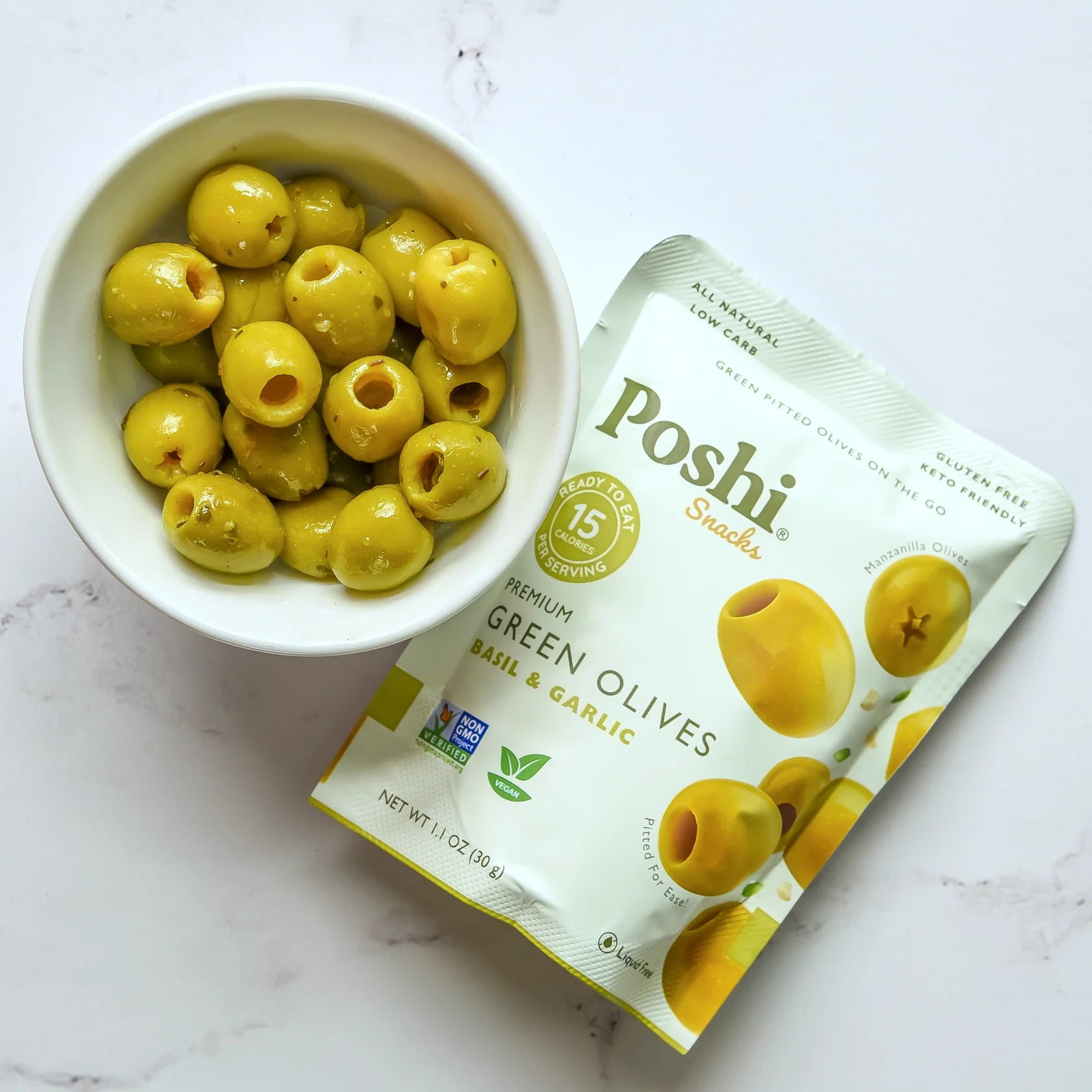 White bowl filled with green olives next to a Poshi Snacks package on a white background